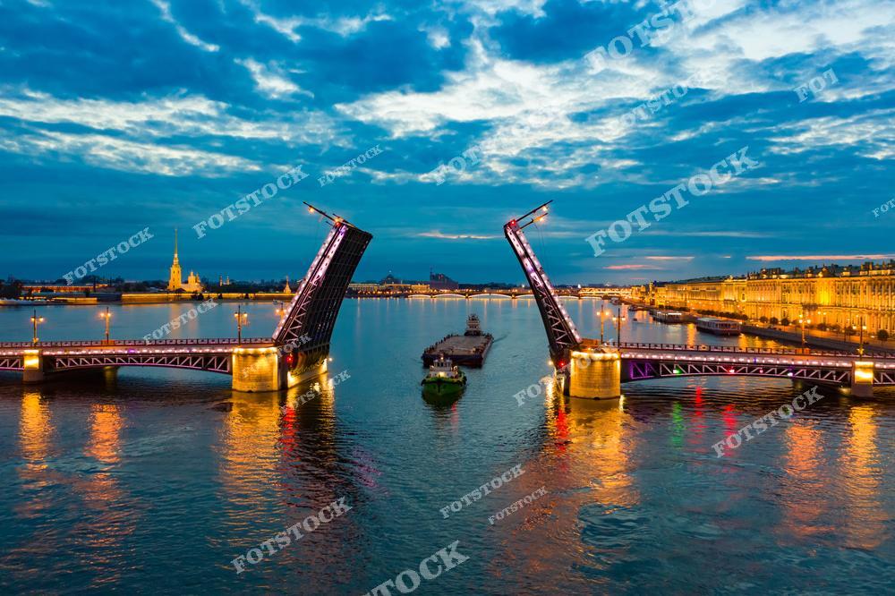 На фотографии изображен разведенный Дворцовый мост в Санкт-Петербурге, освещенный яркими огнями в период белых ночей. На заднем плане можно увидеть Петропавловскую крепость, историческую достопримечательность города, а также фасад Эрмитажа, одного из крупнейших музеев мира. Река Нева освещена отражениями огней города и разводного моста, создавая живописную ночную атмосферу.